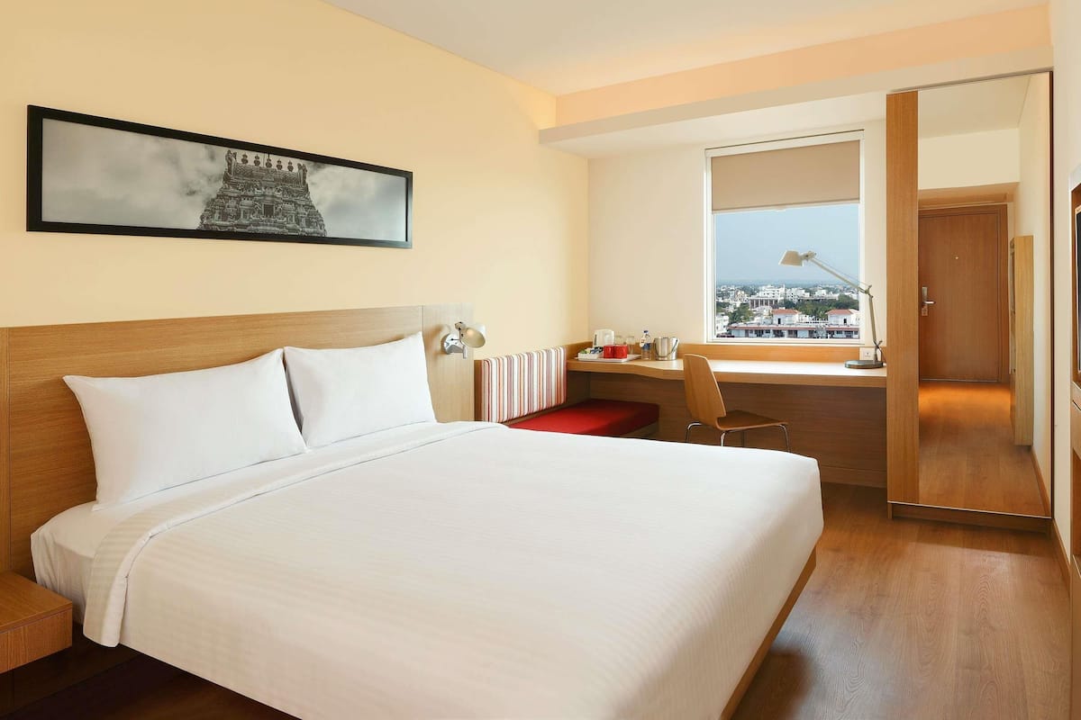 A hotel room with a large bed, a desk with a chair, a window with a view of buildings, and a door.