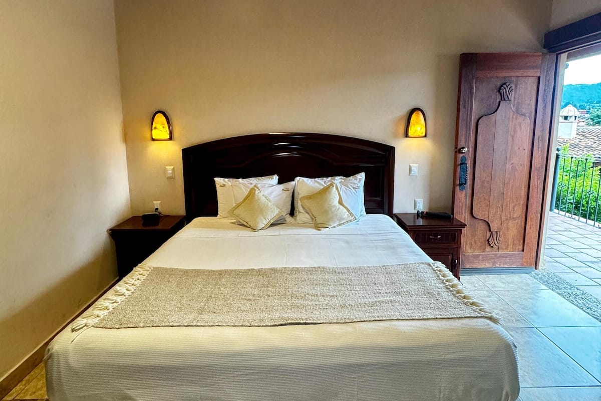 Une chambre avec un plafond en bois, un grand lit recouvert de linge de lit blanc, deux tables de chevet avec des lampes et une porte en bois donnant sur l’extérieur.