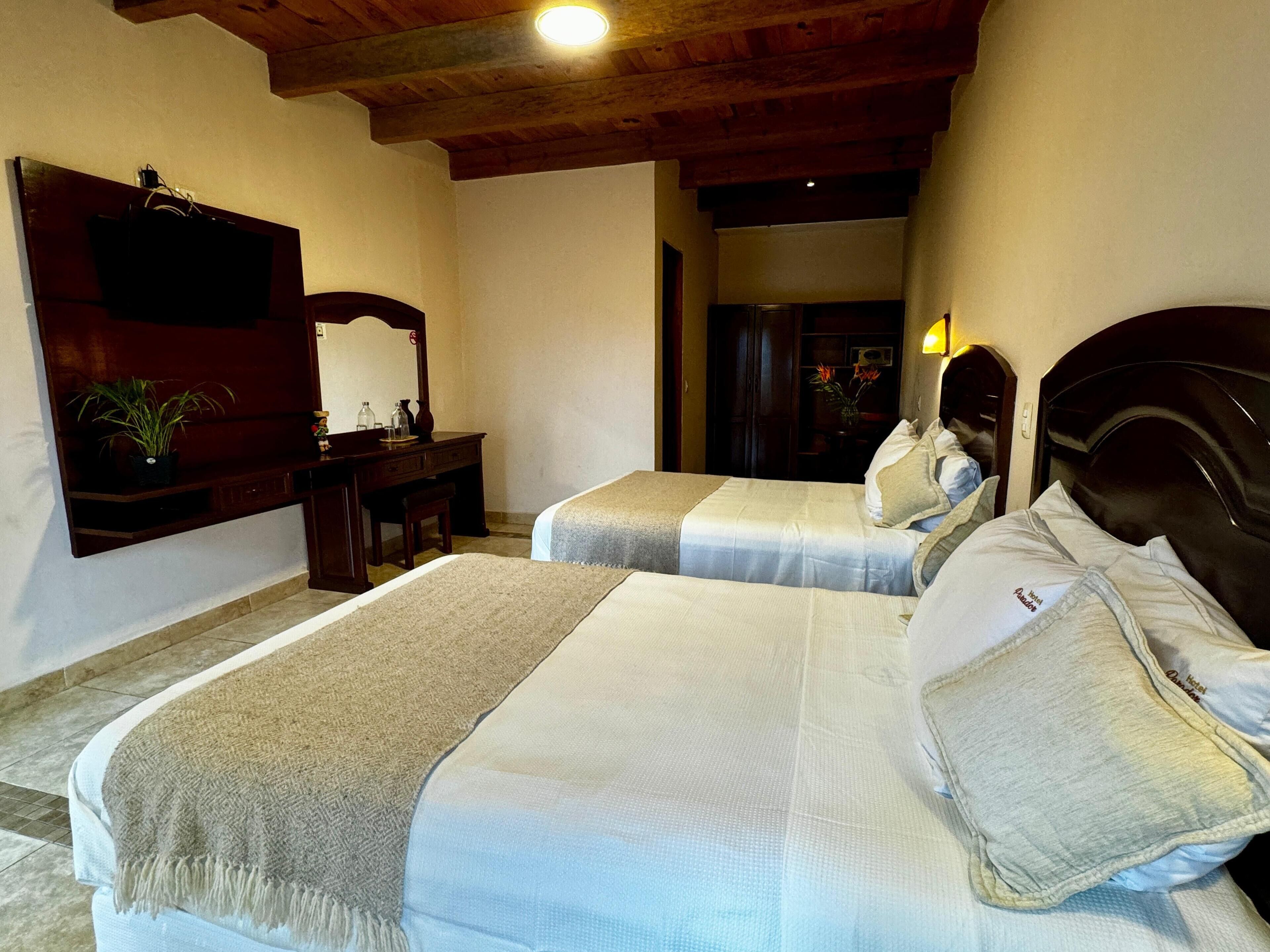 Une chambre d’hôtel avec deux lits, une tête de lit en bois, un bureau avec un miroir et une plante en pot.