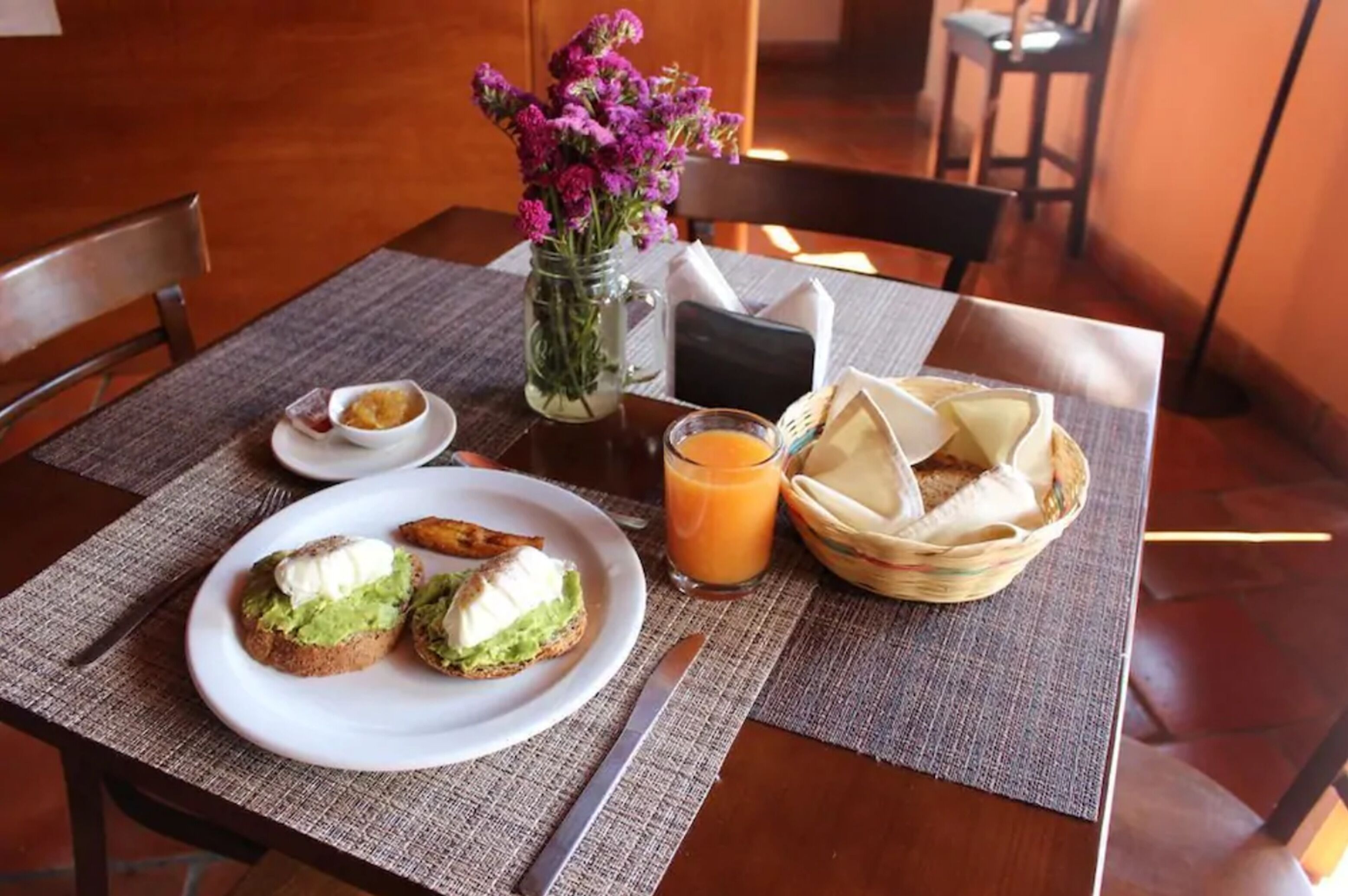 Une table mise pour le petit-déjeuner avec des toasts, un verre de jus d’orange, un petit pot de confiture, un vase de fleurs et un panier de pain.