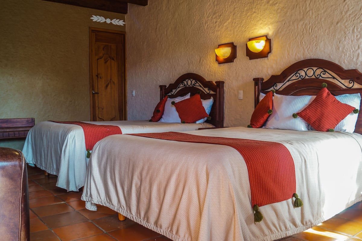 Une chambre d’hôtel avec deux lits, chacun doté d’une tête de lit en bois, de draps blancs et d’oreillers rouges. On y trouve des lampes fixées au mur et une porte en bois.