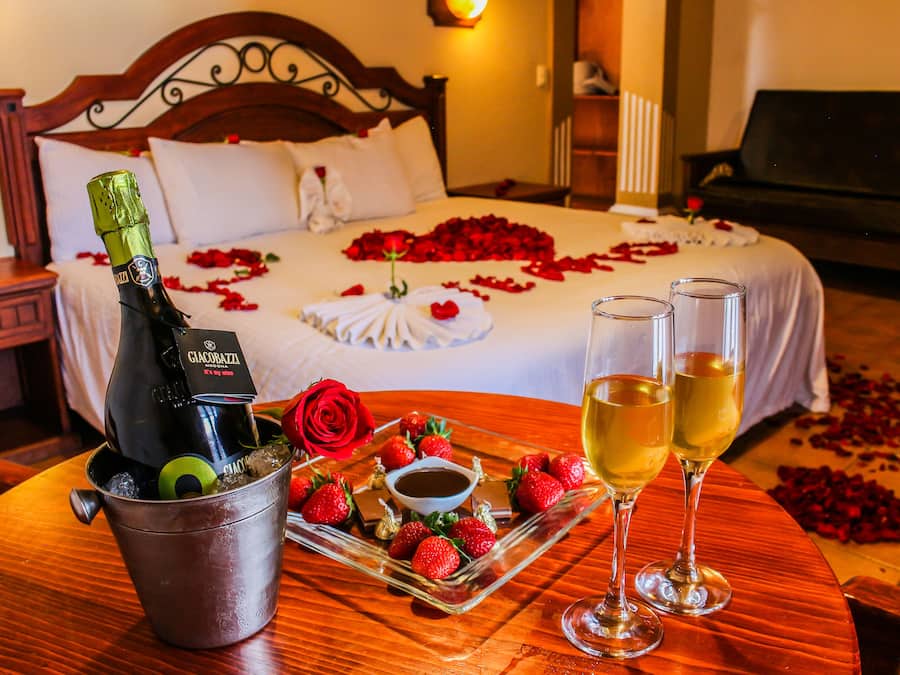 A hotel room with a bed, a bedside table with a bottle of wine, strawberries, and a tray of food.