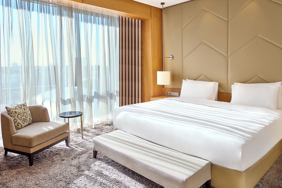 Habitación de hotel con una cama grande, un sofá, una mesita y vistas a la ciudad a través de cortinas translúcidas.