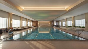 Une vaste piscine intérieure à l’eau d’un bleu limpide, entourée d’espaces de détente avec des canapés et des fauteuils, et dotée de grandes fenêtres de part et d’autre.