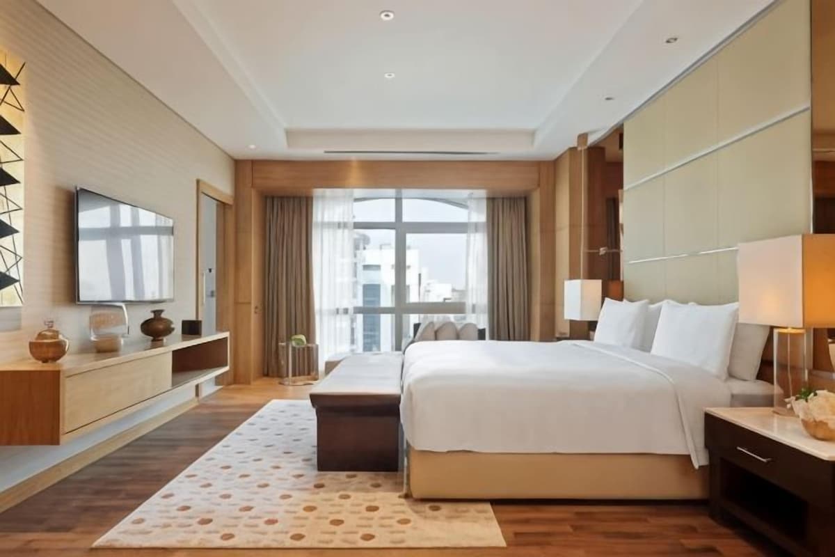 A modern hotel room with a large bed, a flat-screen TV, a desk, and a large window.