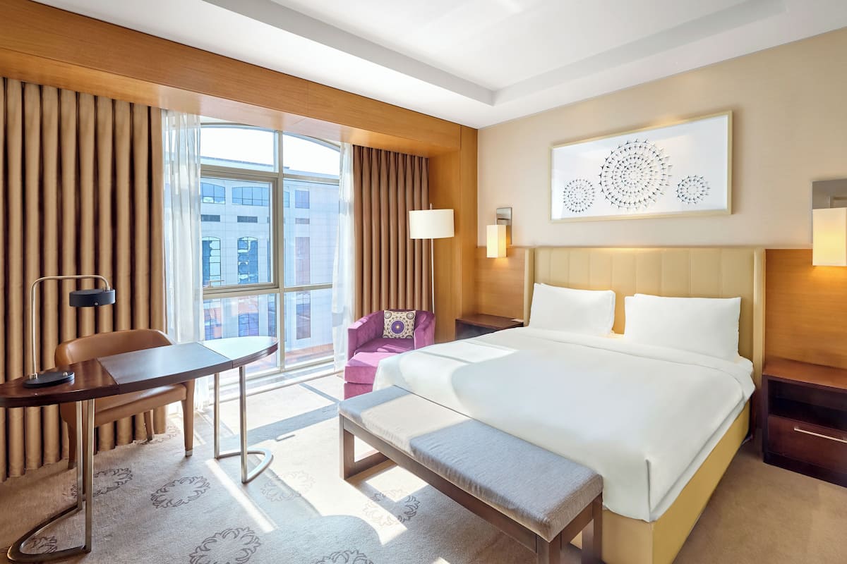 Habitación de hotel con una cama grande, un escritorio, una silla, un banco y vistas a la ciudad desde la ventana.