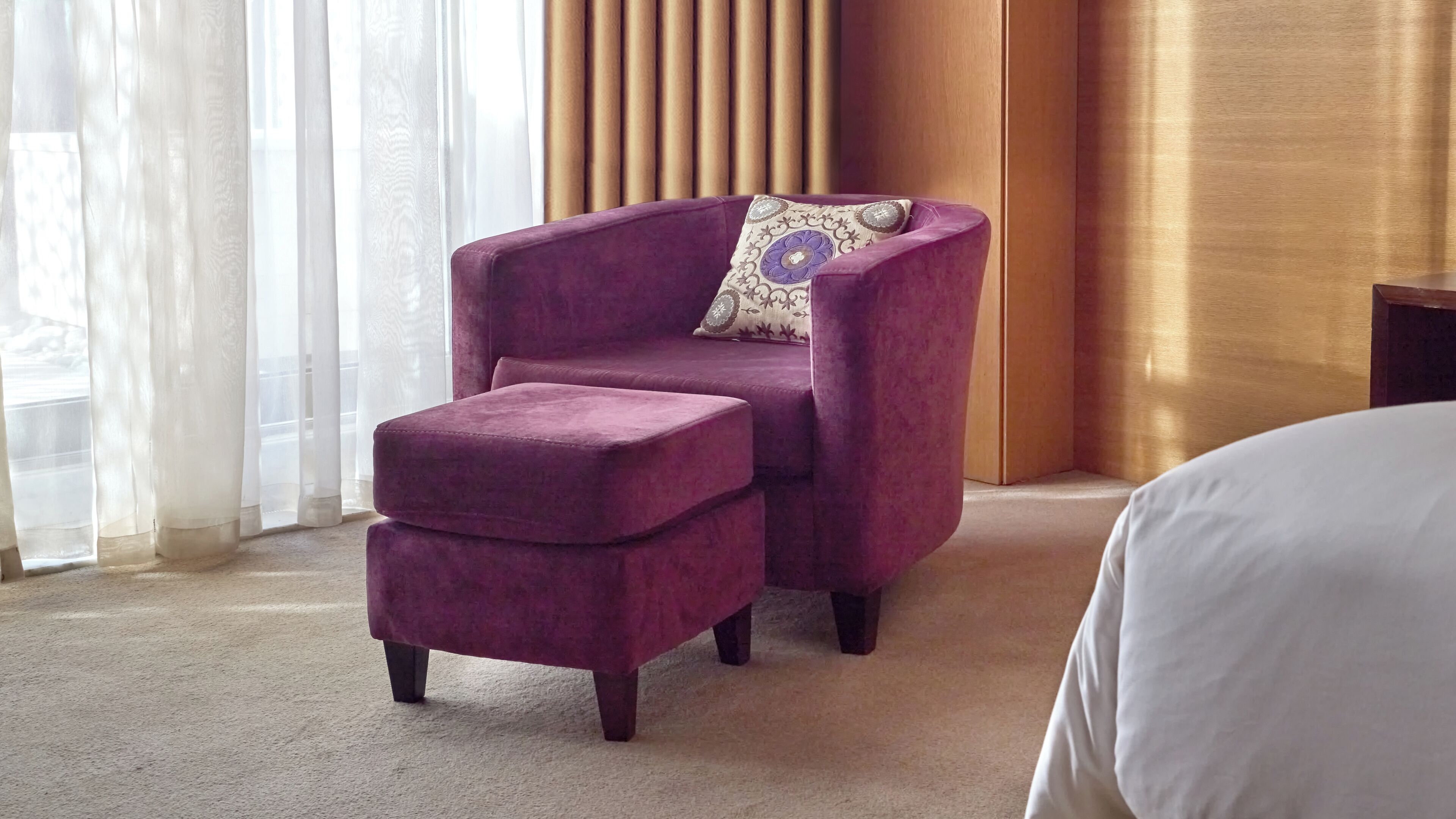 Una habitación con un sillón púrpura y un reposapiés, una cama con ropa de cama blanca, y una ventana con cortinas translúcidas.