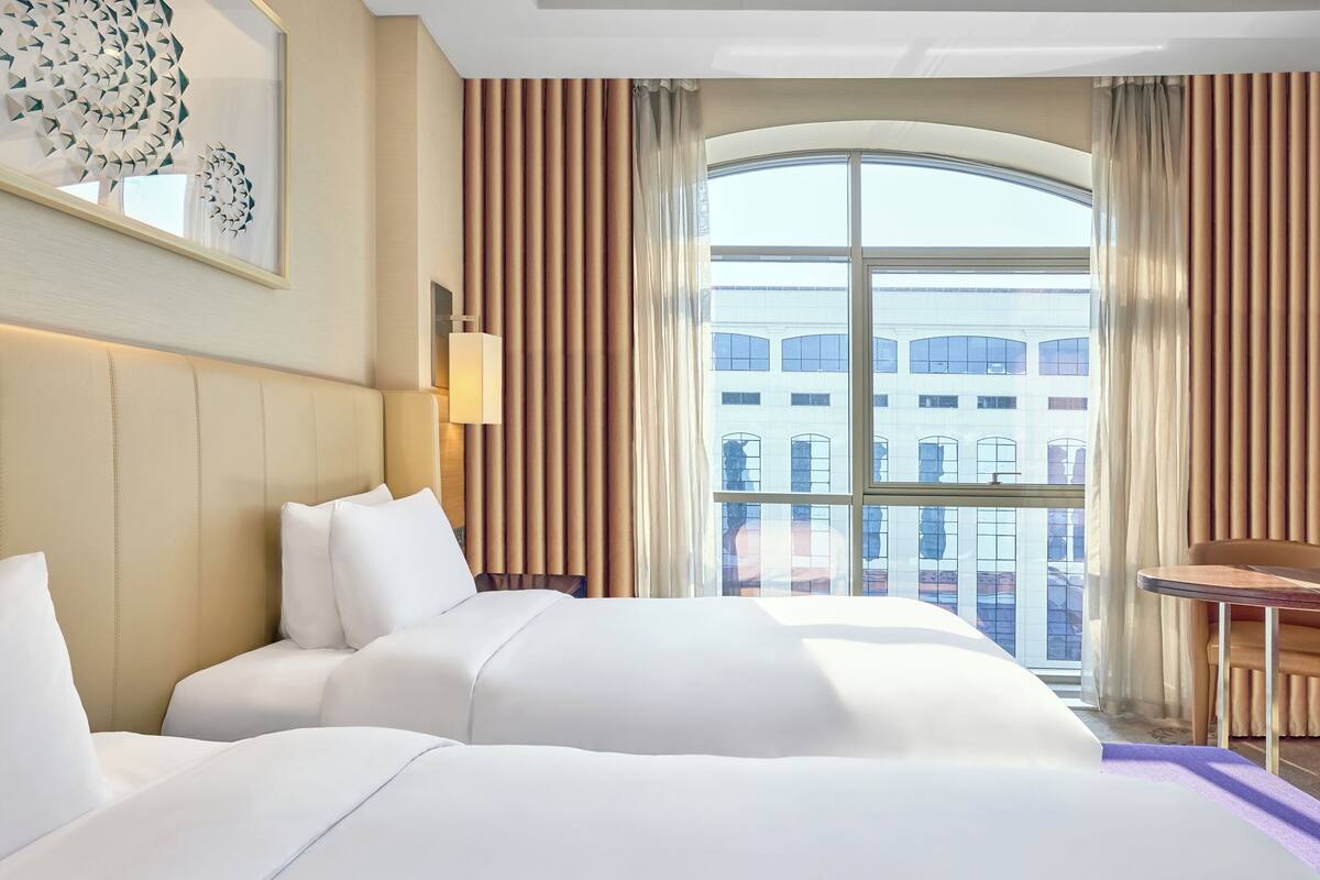 Habitación de hotel con dos camas, un ventanal con cortinas, un escritorio y vistas a edificios.