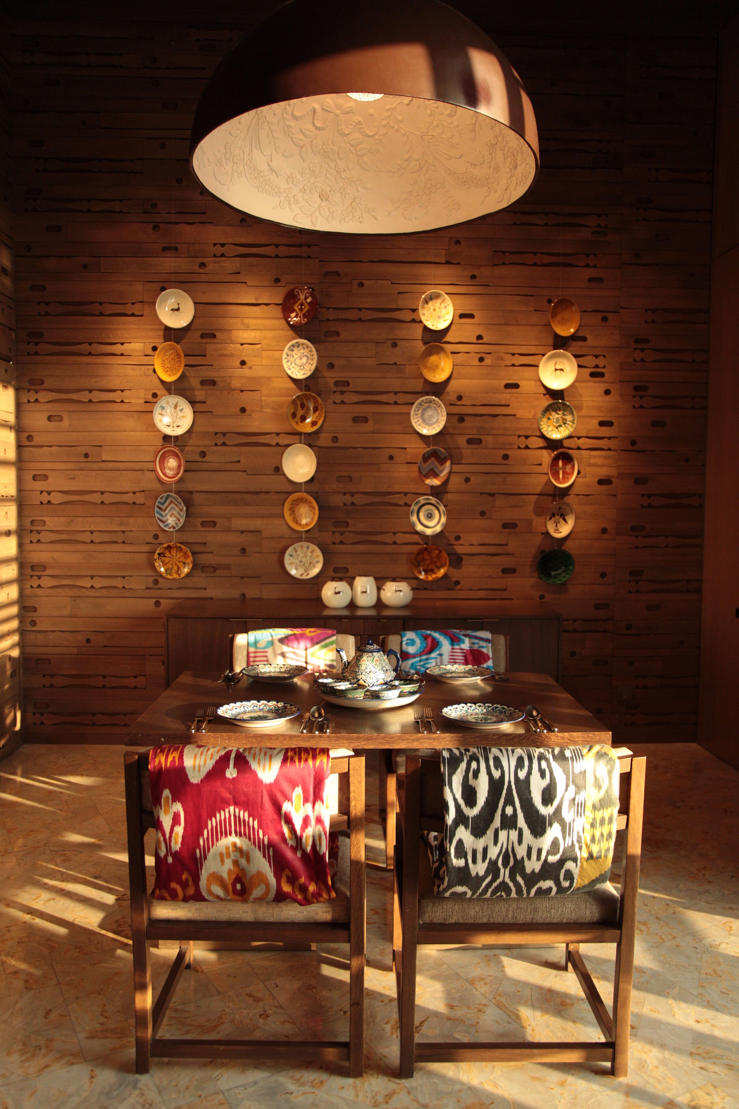 A dining area with a wooden wall, a large hanging lamp, and a table set with plates and cutlery.
