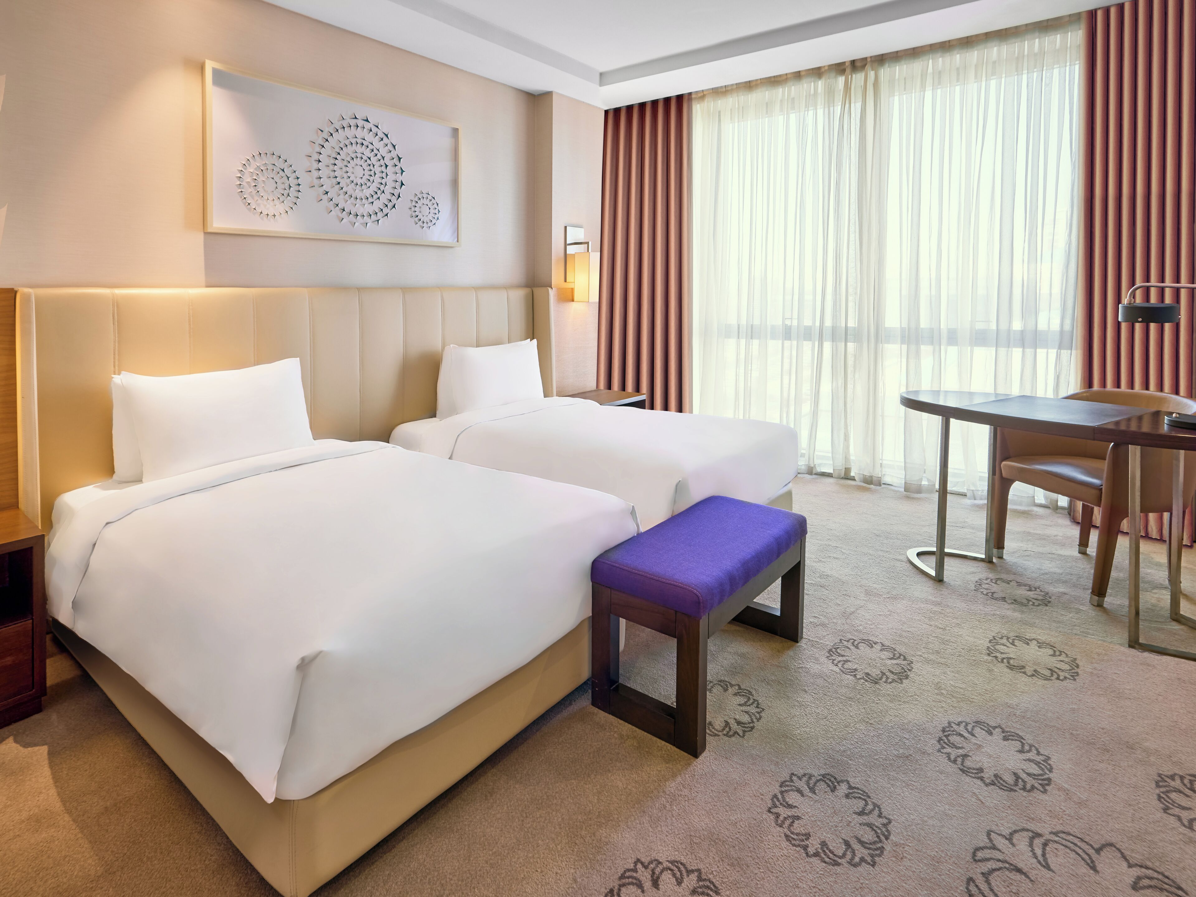 A hotel room with two beds, a small table, a bench, a wall-mounted artwork, and a large window with curtains.