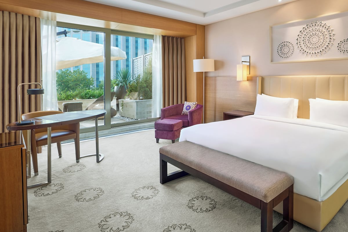 Habitación de hotel con una cama grande, un escritorio, una silla, un banco, una lámpara y vistas al exterior con vegetación.