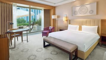 Une chambre d’hôtel avec un grand lit, un bureau, une chaise, un banc, une lampe et une vue sur la verdure environnante.