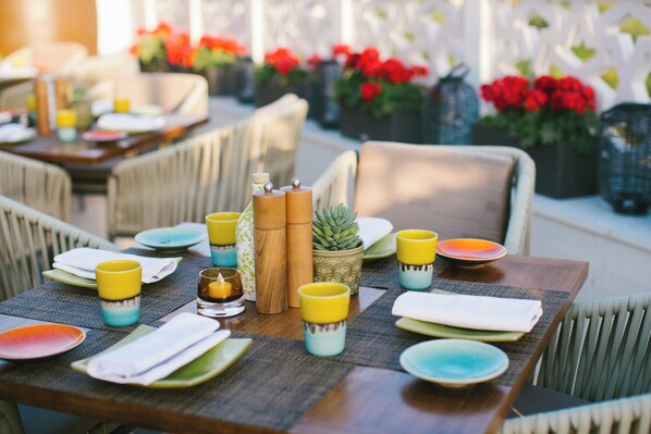 Une table à manger dressée avec des assiettes, des serviettes et des tasses colorées, entourée de chaises en osier et de plantes en pot.