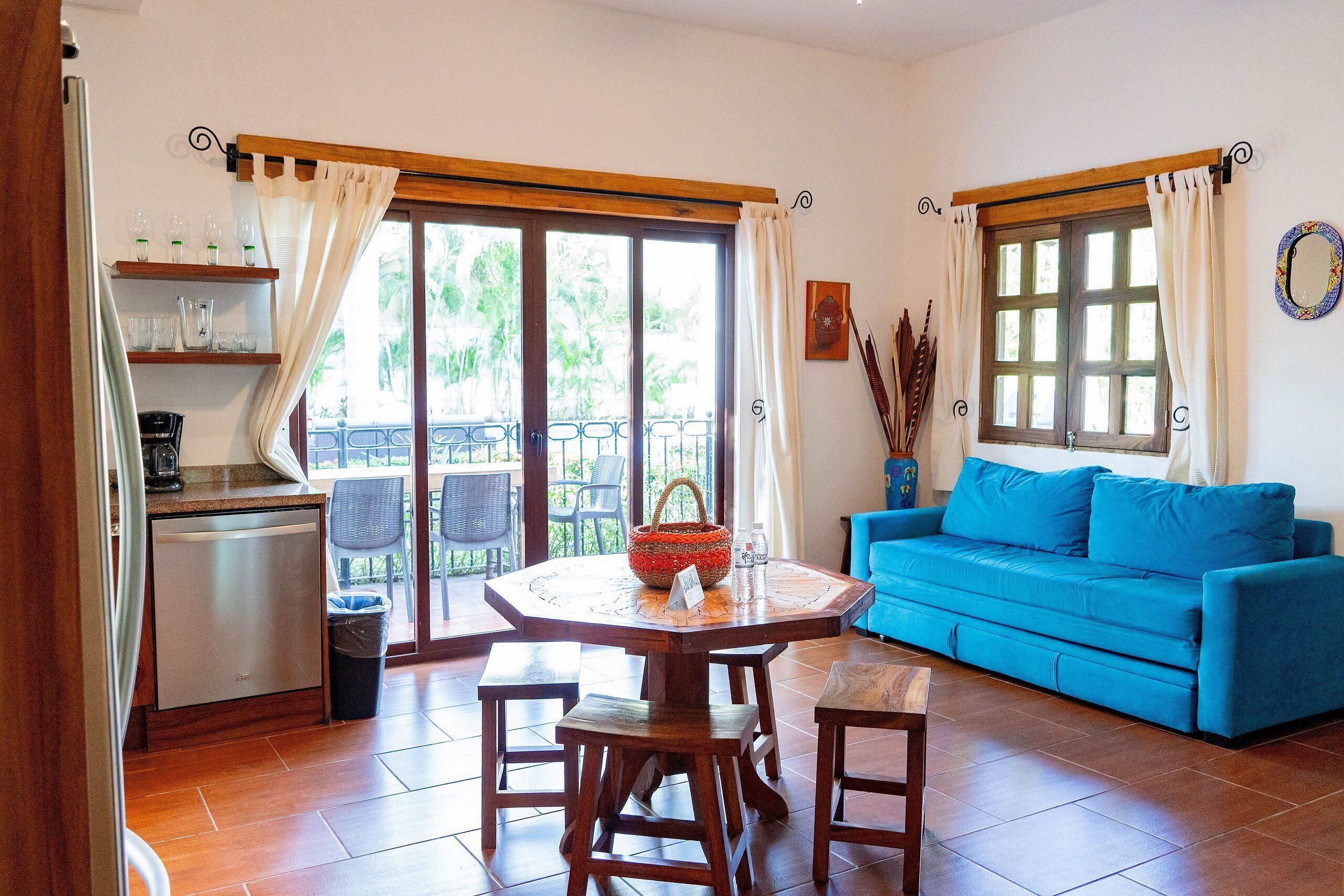 Een knus woongedeelte met een kitchenette, een eettafel, een blauwe bank en een balkon met uitzicht op groen.