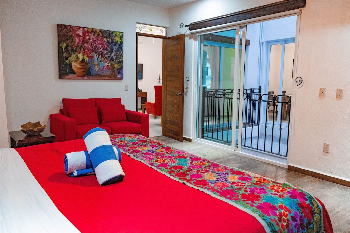 Ein Hotelzimmer mit einem Bett, einem roten Sofa, einem farbenfrohen Teppich, einem Bild an der Wand und einer Glastür zum Balkon.