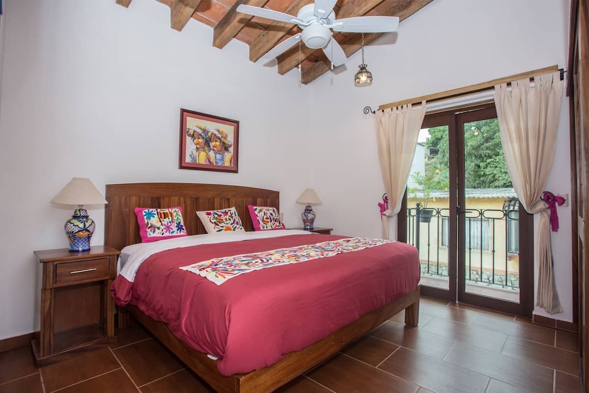 Ein Schlafzimmer mit einem hölzernen Bett, zwei Nachttischen mit Lampen, einem Deckenventilator, einem Bild an der Wand und einem Balkon mit Vorhängen.