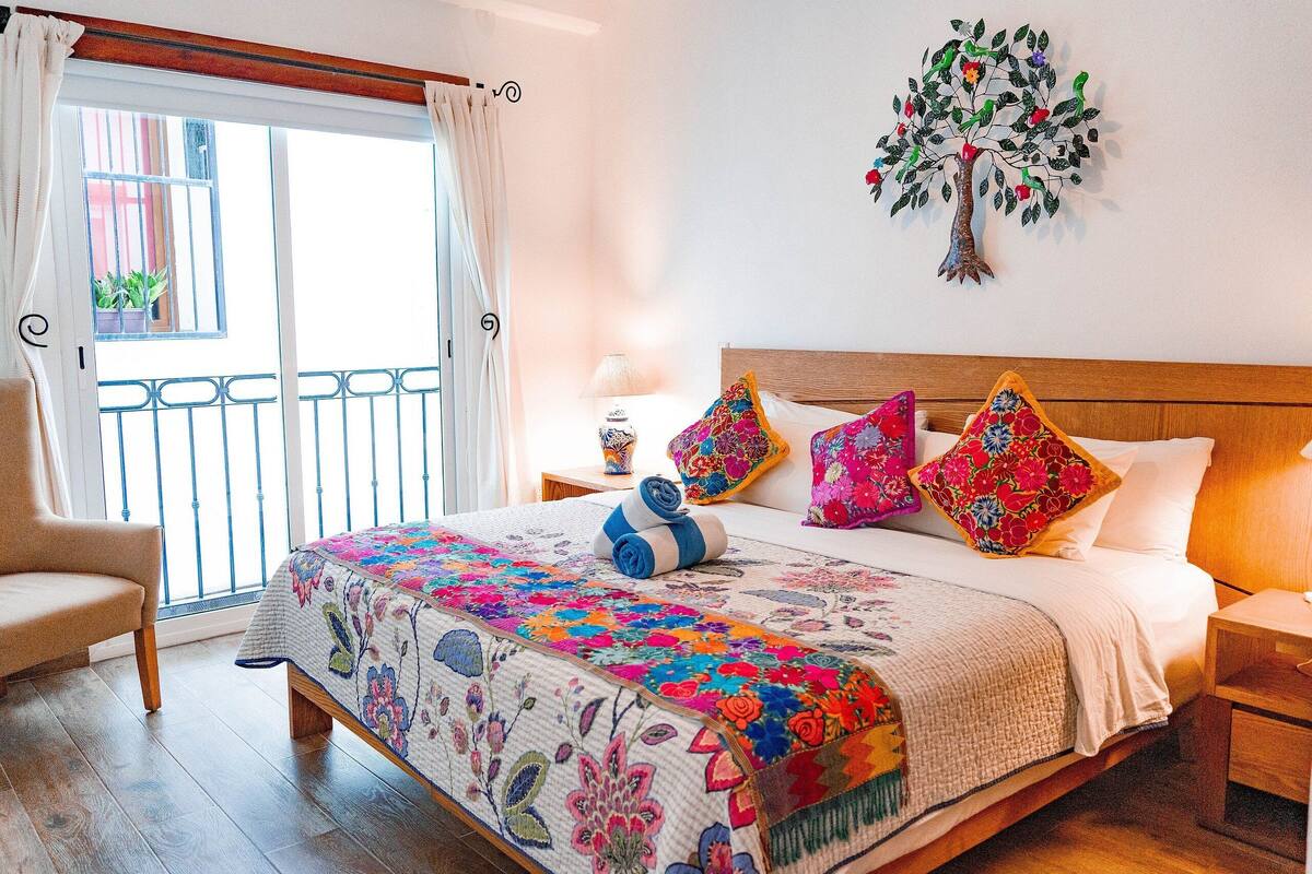 Ein Schlafzimmer mit einem Holzbett, farbenfroher Blumenbettwäsche, Wandkunst mit einem Baumdekor und einem Balkon mit Blick, sowie einem Sessel.
