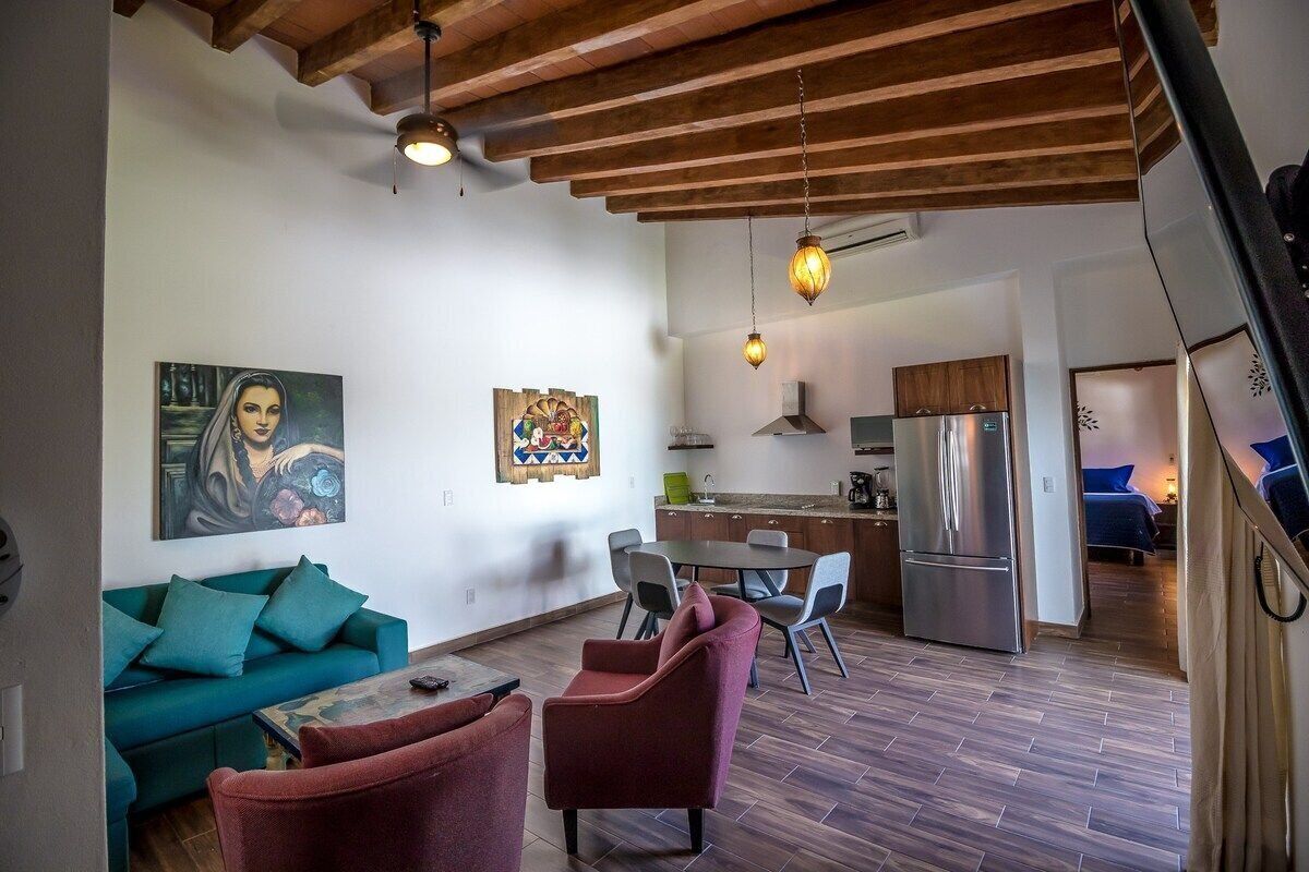 A modern living room with a sofa, armchairs, and a dining table. There is a kitchen in the background with a refrigerator and cabinets.