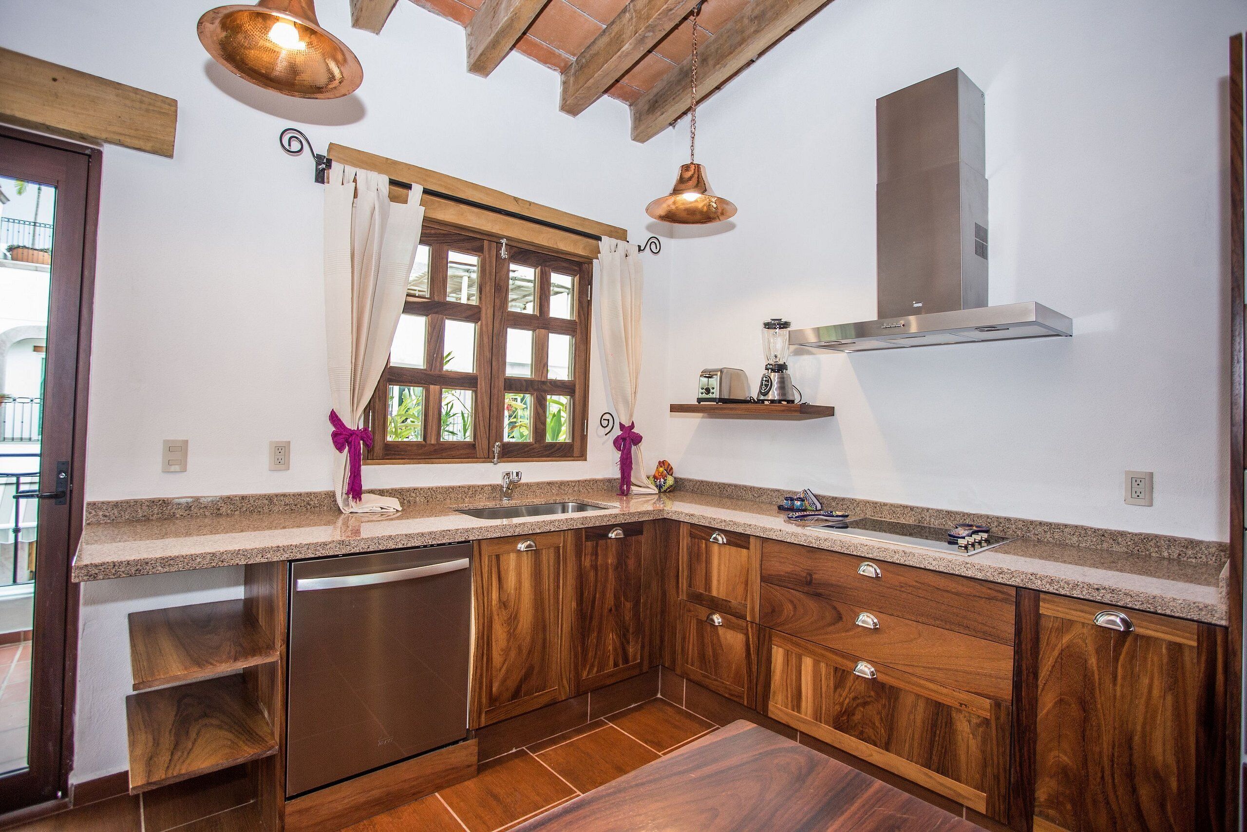 A kitchen with wooden cabinets, a stainless steel dishwasher, a window with curtains, and pendant lighting.