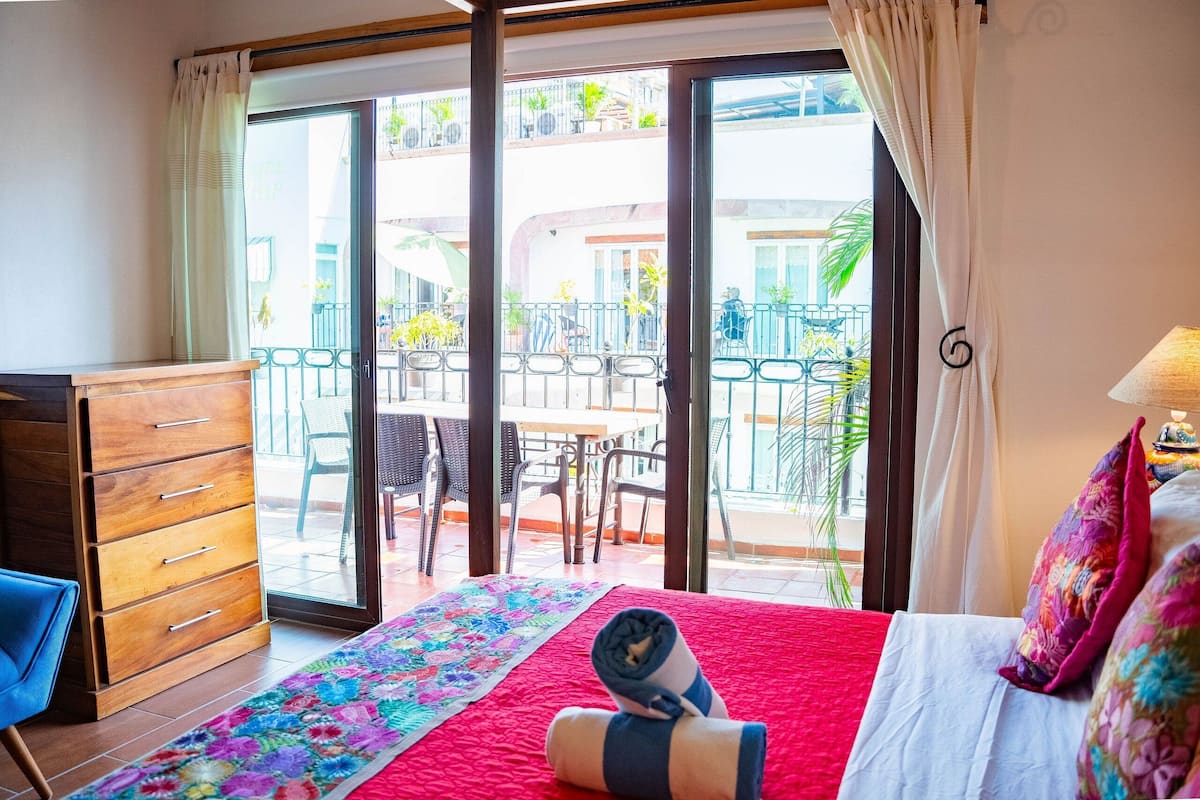 Ein Hotelzimmer mit Bett, Schminktisch, Fenster mit Vorhängen, Balkon mit Stühlen und Tischen sowie Blick nach draußen.