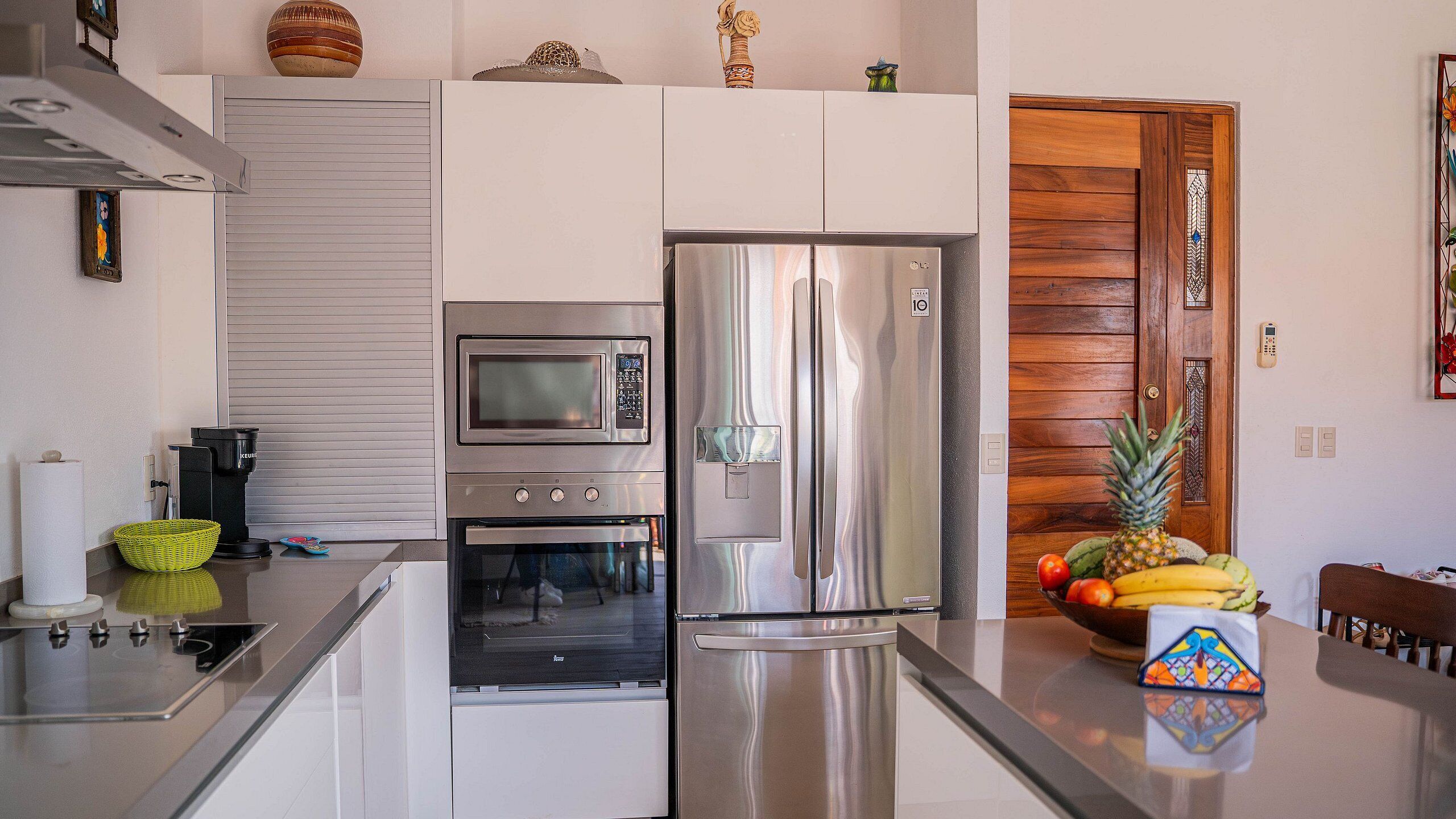 A modern kitchen with a stainless steel refrigerator, built-in oven, and microwave. There is a wooden door with a decorative glass panel, and a fruit bowl on the counter.