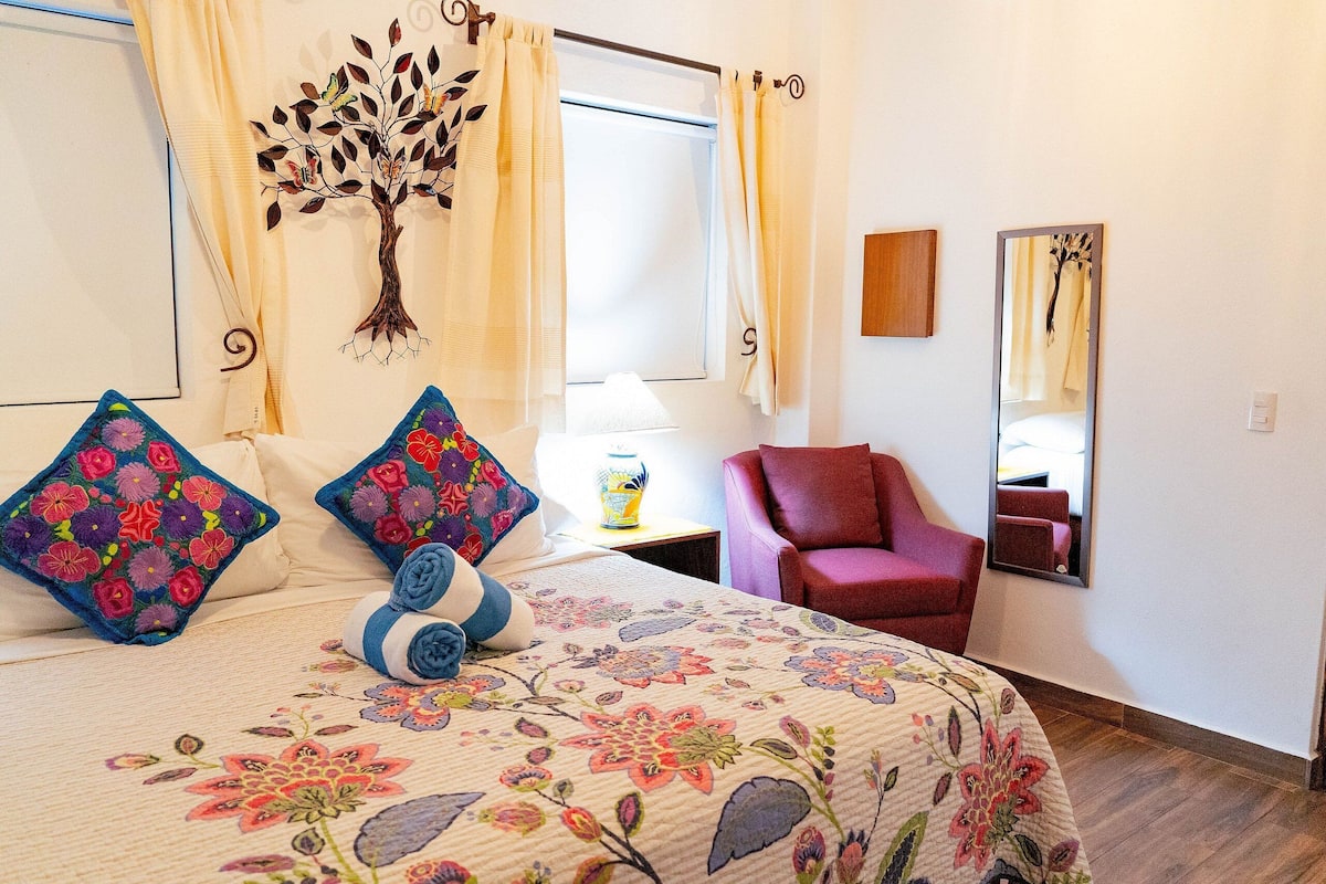 Ein Schlafzimmer mit floralem Bettüberwurf, einer Wanddekoration mit Bäumen, einem roten Sessel und einem Spiegel an der Wand.