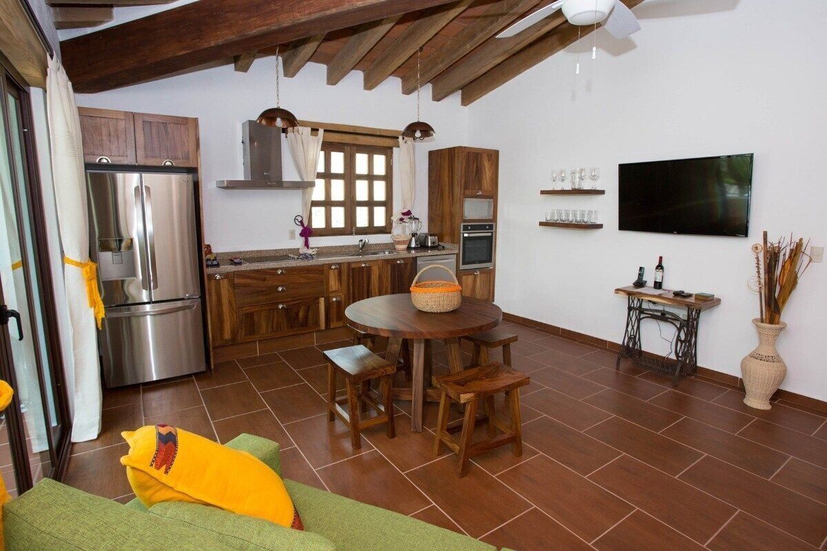 A compact kitchen with modern appliances, a dining area with wooden furniture, and a flat-screen TV mounted on the wall.