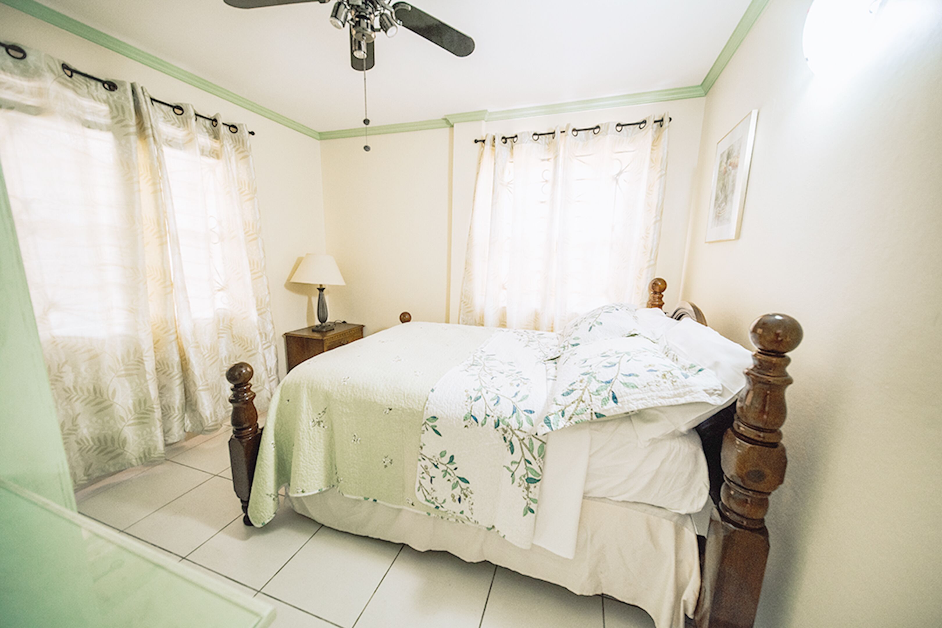 Une chambre à coucher avec un lit, des rideaux, une table de chevet, une lampe et un ventilateur de plafond.
