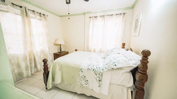 Une chambre à coucher avec un lit, des rideaux, une table de chevet, une lampe et un ventilateur de plafond.