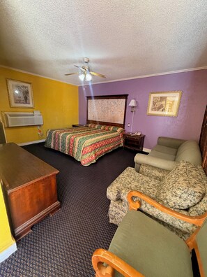 A hotel room with a bed, a desk, a chair, a ceiling fan, and two framed pictures on the walls.