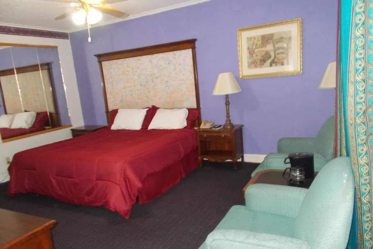 Quarto de hotel com uma cama grande, uma mesinha de cabeceira com luminária, uma cadeira, uma cafeteira e um quadro pendurado na parede.