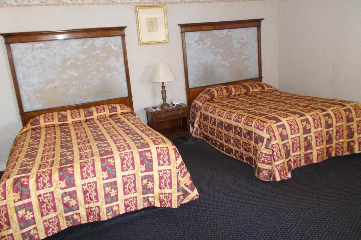 Quarto com duas camas, cada uma com cabeceira de madeira e colcha estampada. Há uma mesinha de cabeceira com abajur entre as camas.