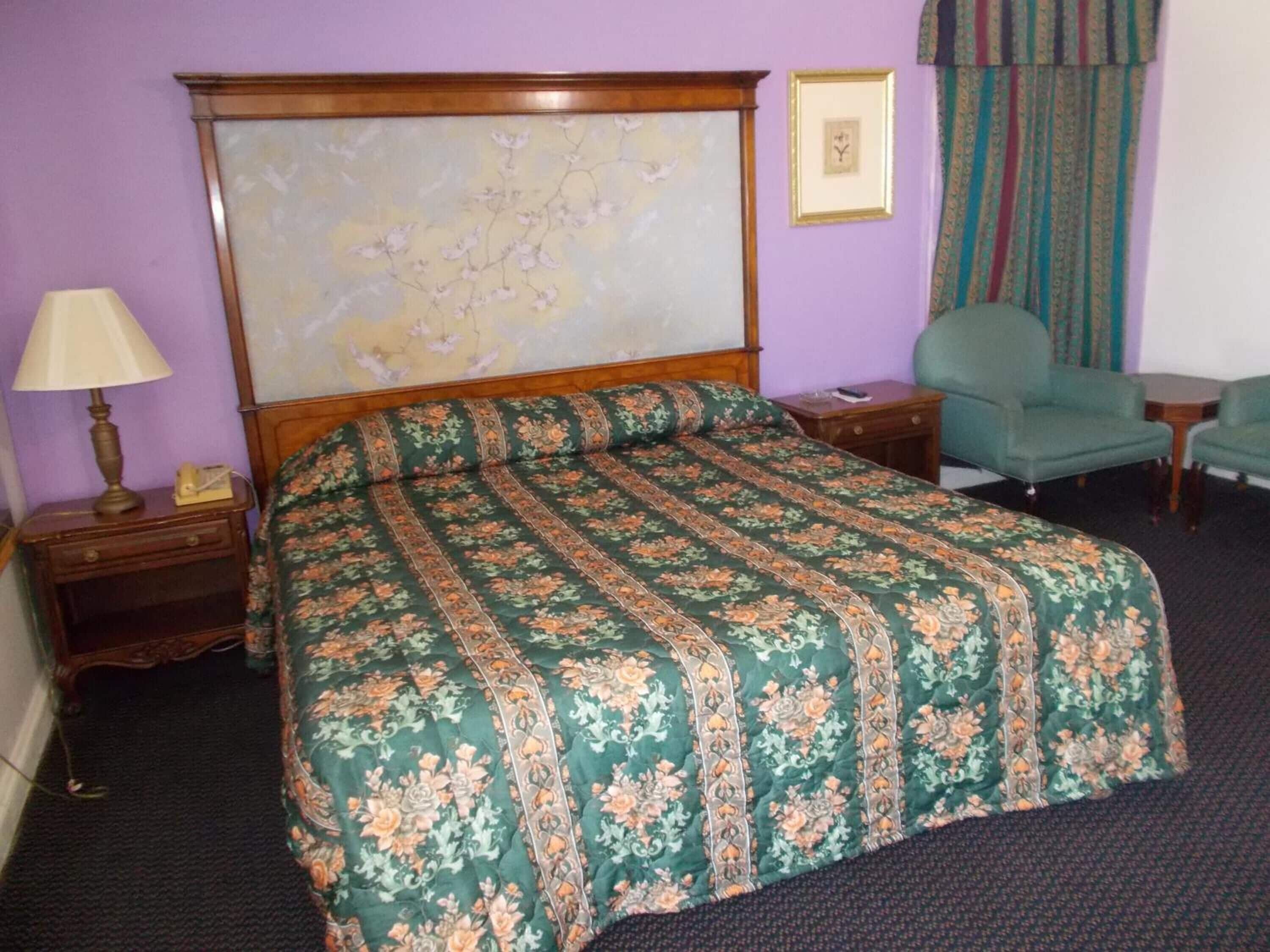Een keurig opgemaakt bed met een bloemenbedekker, een houten hoofdbord, een nachtkastje met een lamp en een groene fauteuil.