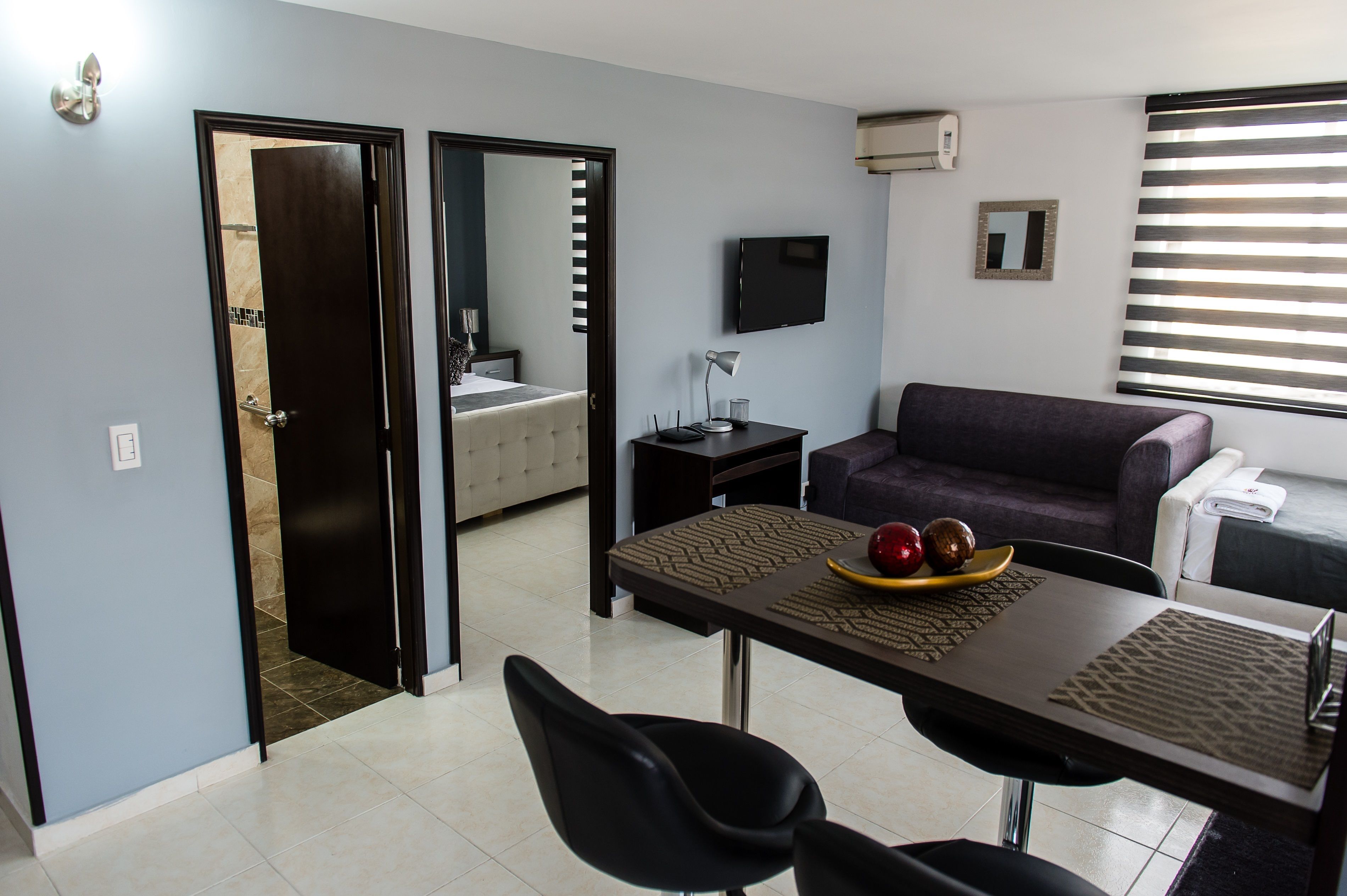 A modern hotel room with a dining area, a sofa, a TV, and a bathroom visible through an open door.