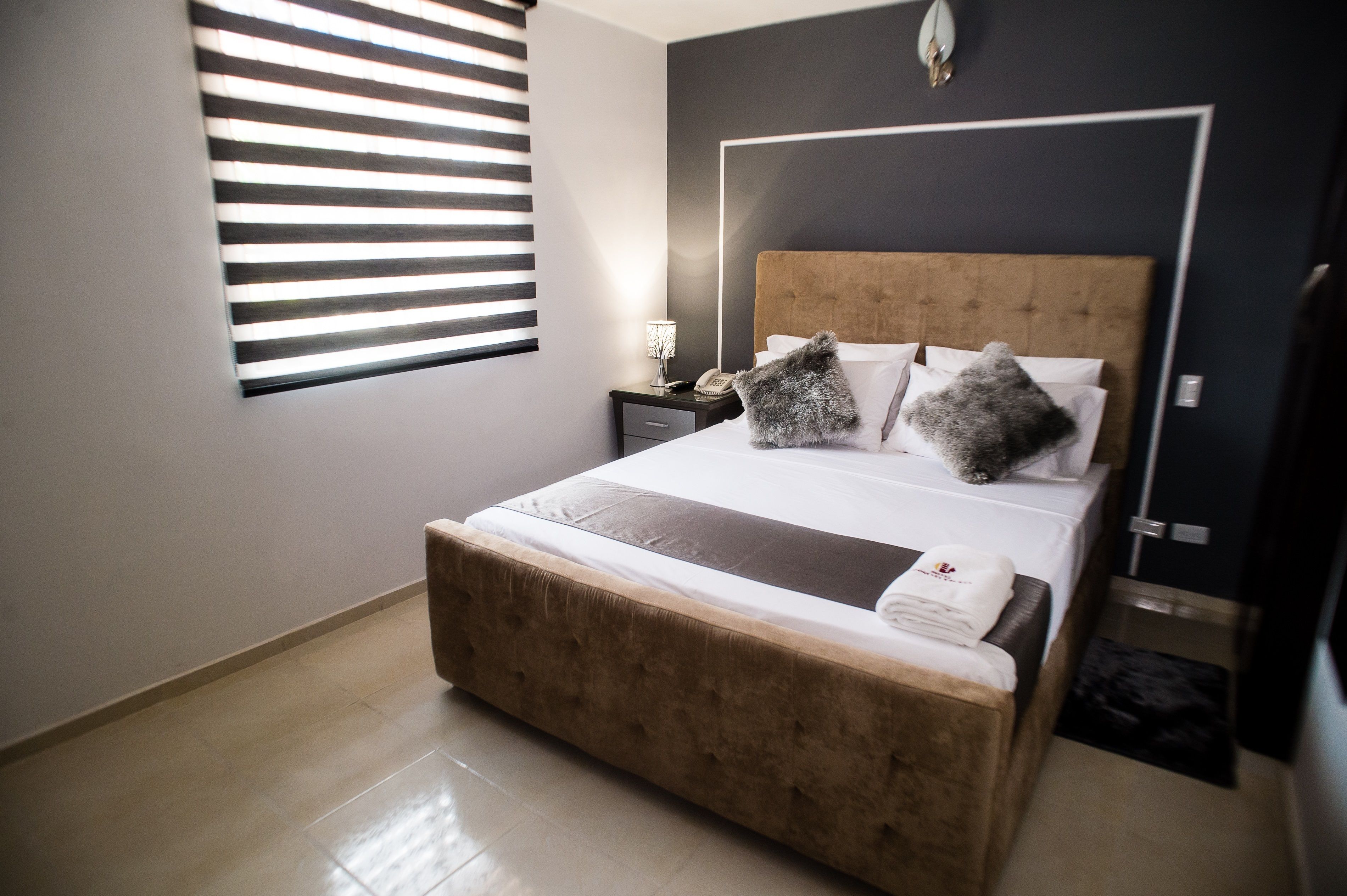 A neatly made bed with a brown headboard, a bedside table with a lamp, and a window with blinds.
