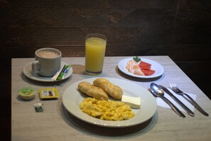 Une table dressée pour le petit-déjeuner avec une tasse de café, un verre de jus d’orange, des croissants, des œufs brouillés, une tranche de fromage et une assiette avec une tomate et une orange.