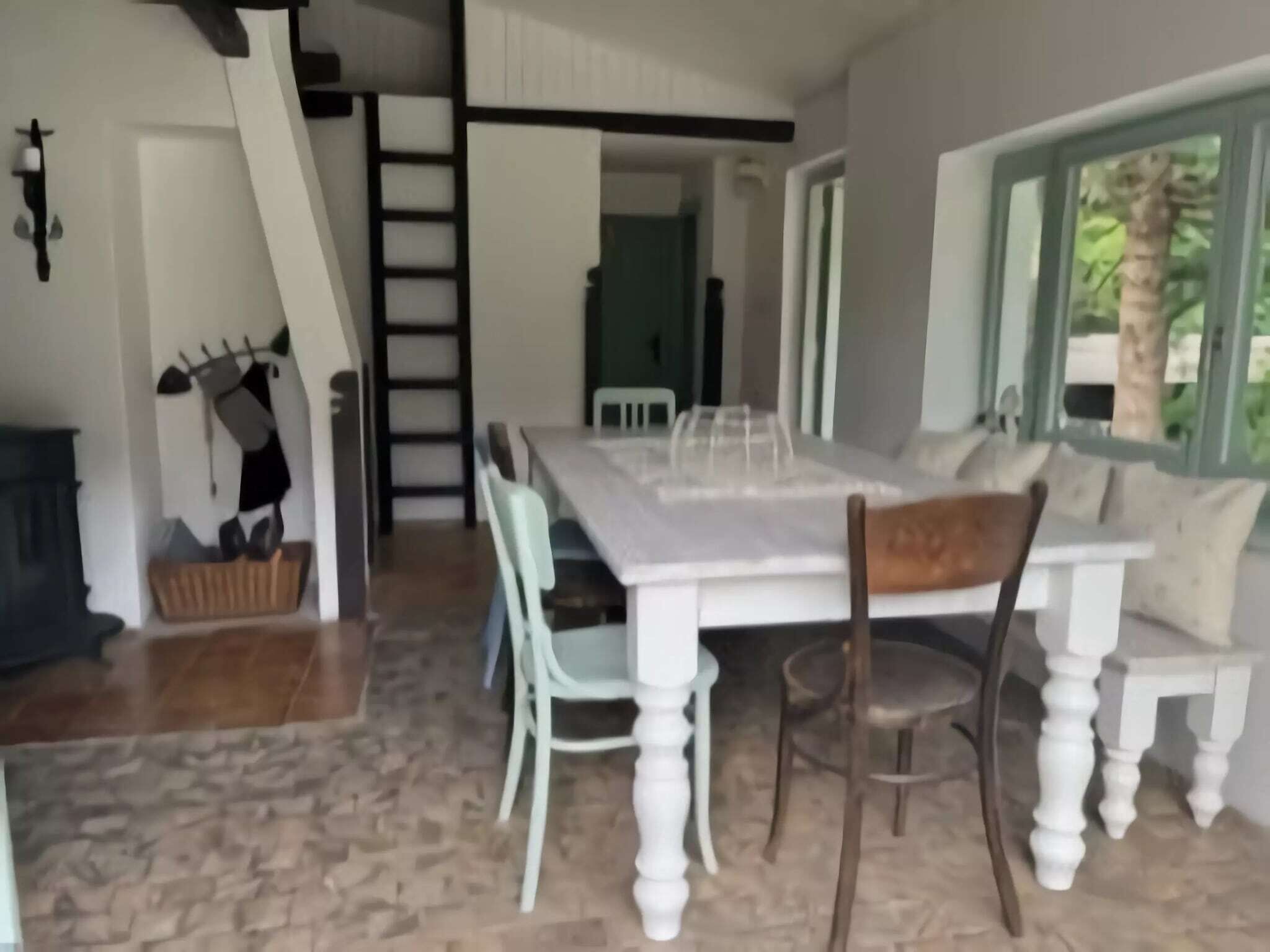 A dining area with a white table and chairs, a wooden bench, a ladder, and a view of greenery outside.