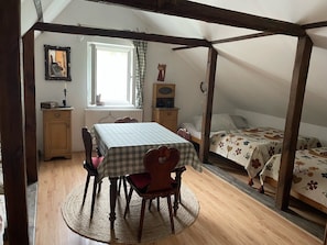 Una habitación con una mesa de comedor, sillas, una cama, una ventana, un radiador, un mueble pequeño y un cuadro en la pared.