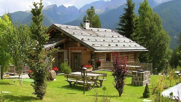 Uma cabana de tronco com telhado inclinado, rodeada por um gramado bem cuidado e uma cerca de madeira.