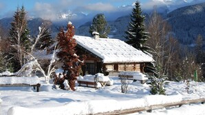Uma cabana de tronco com telhado coberto de neve, rodeada por árvores carregadas de neve e montanhas ao fundo.