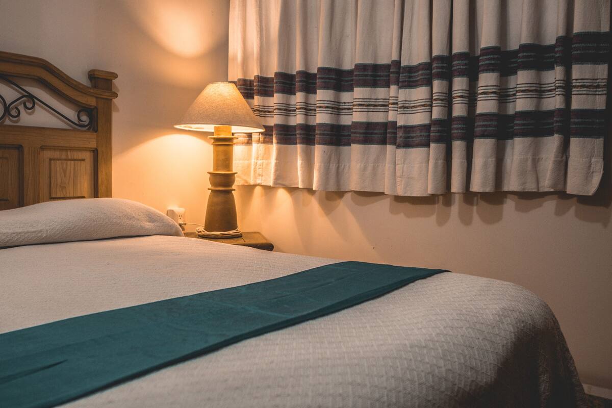 A neatly made bed with a green throw blanket, a wooden headboard with decorative carvings, a bedside lamp, and striped curtains.