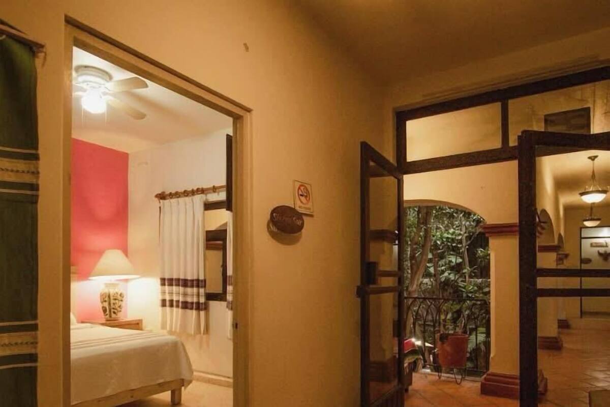 A hotel room with a bed, a nightstand, a lamp, and a view of a balcony with plants.