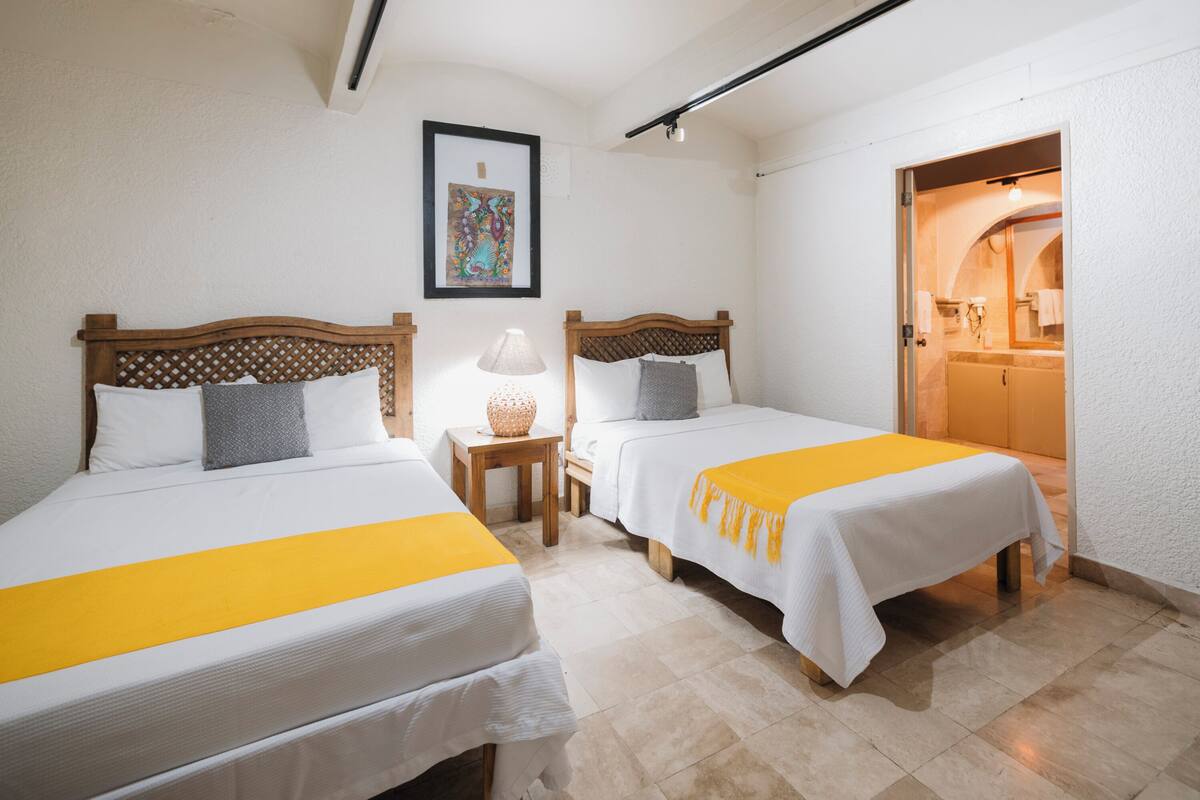 A hotel room with two beds, a small table, a lamp, and a bathroom visible through an open door.