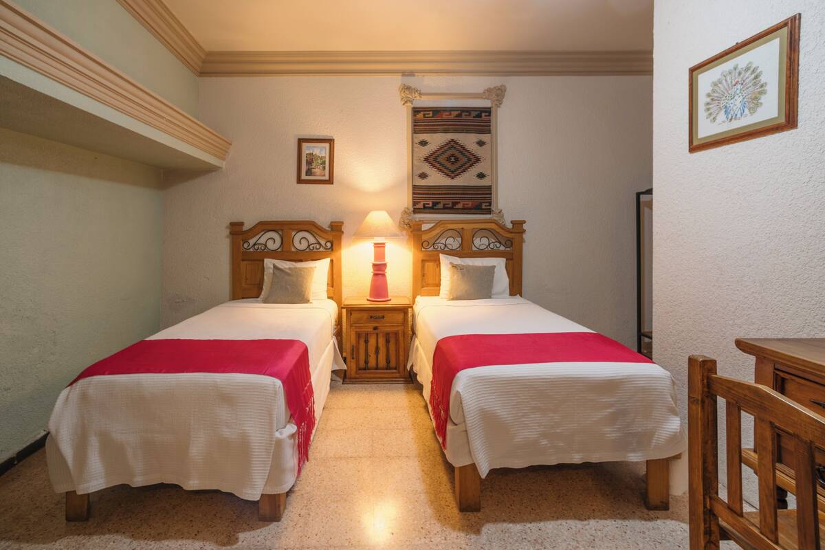 A room with two single beds, a nightstand with a lamp, a wooden chair, and a decorative wall hanging.