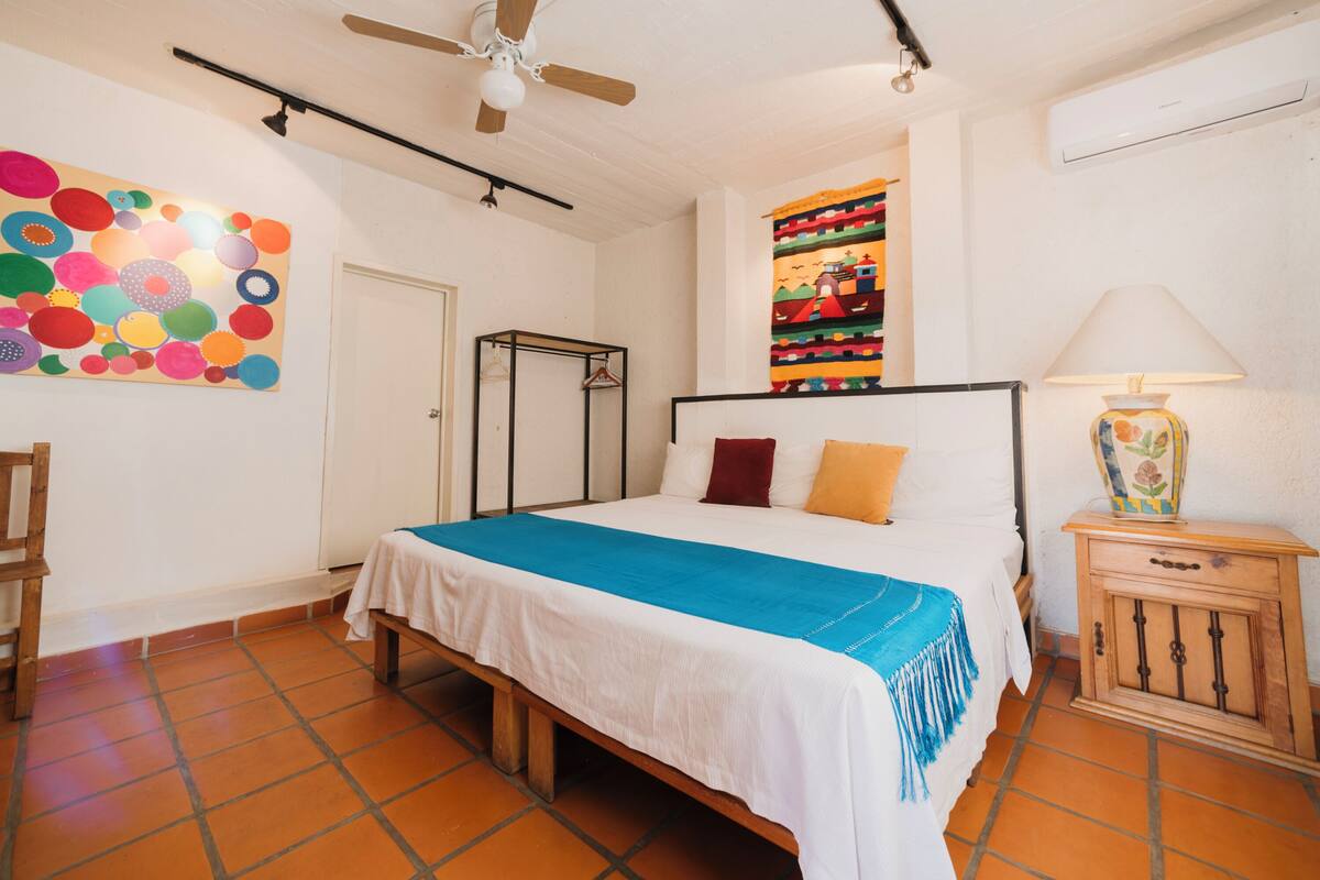 A bedroom with a bed, a ceiling fan, a colorful wall hanging, a wooden cabinet, and a tiled floor.