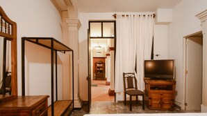 A hotel room with a bed, a wooden dresser, a television on a wooden cabinet, a chair, and a door leading to a hallway with a staircase. - Hotel Casa Guivá (Oaxaca)