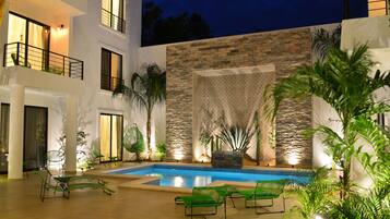 A modern outdoor pool area with lounge chairs, a stone wall, and a view of a multi-story building at night.
