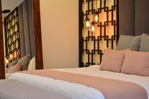 A neatly made bed with a white and peach-colored bedspread, a wooden headboard, and a decorative wall panel with hanging lights.