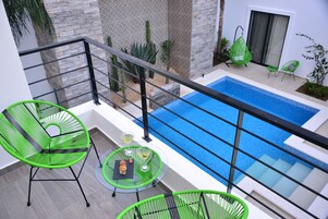 Un balcón con piscina, sillas verdes y una mesa con bebidas.