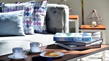 Una sala acogedora con un sofá, almohadas, una mesita de centro con un libro y galletas, y una mesita auxiliar con una vela.